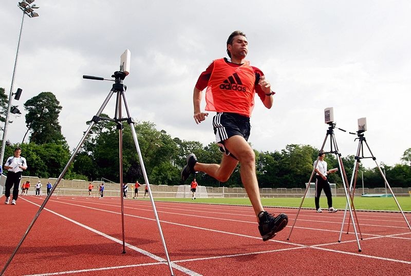 Athlete in a red Adidas shirt running on a track with timing devices on tripods.