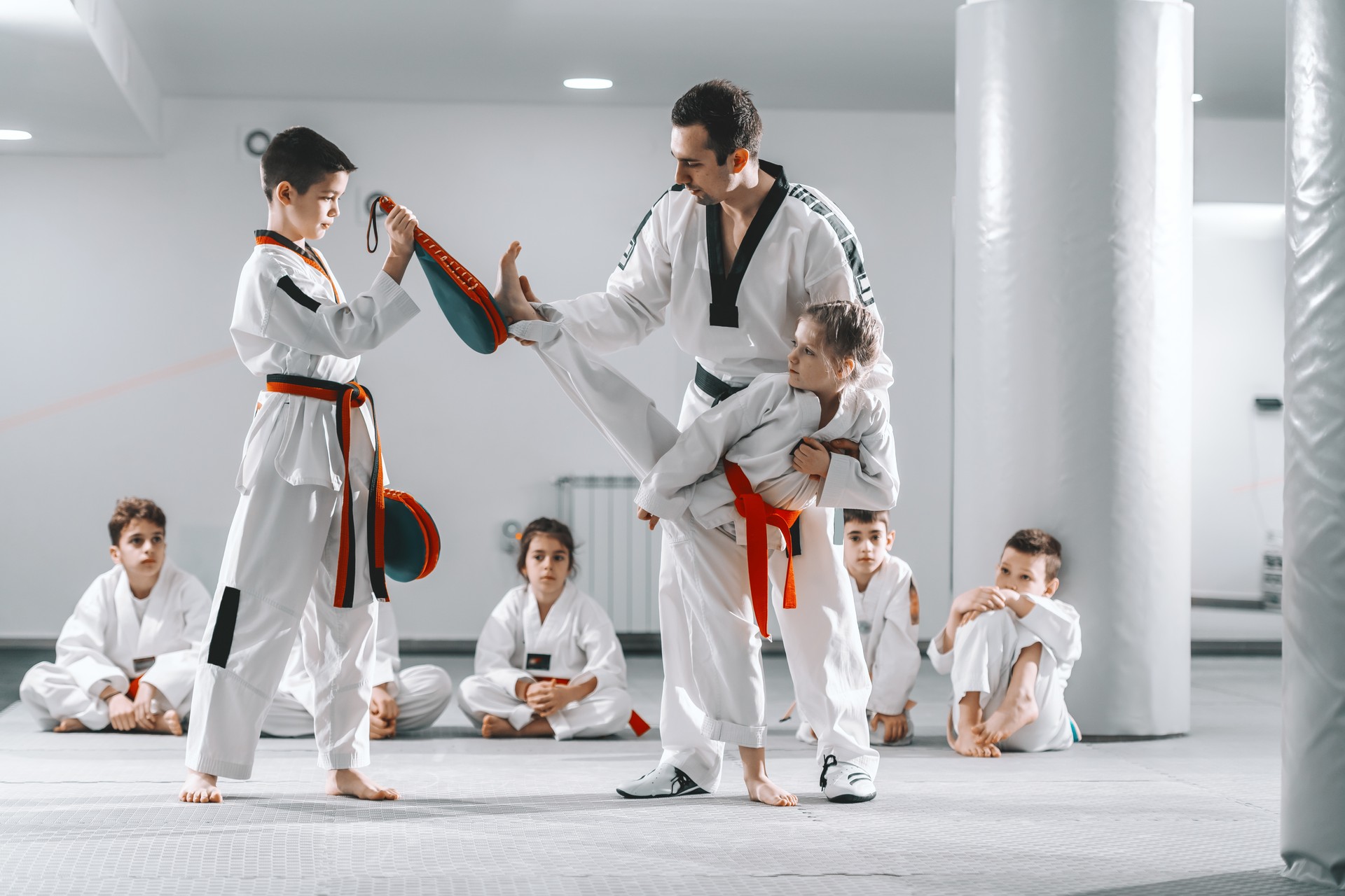 Trainer showing to young girl how to kick kick target. Boy holding target. Tekwondo training concept.